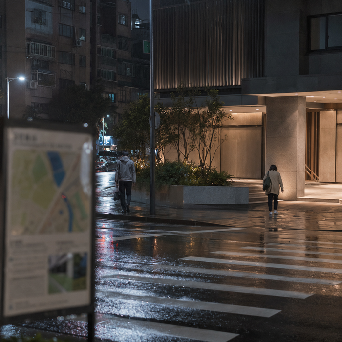 雨夜城市路口與現代建築前的匿名行人,呈現定點茶被視為觀光賣點時所引發的公共治理與道德爭議。